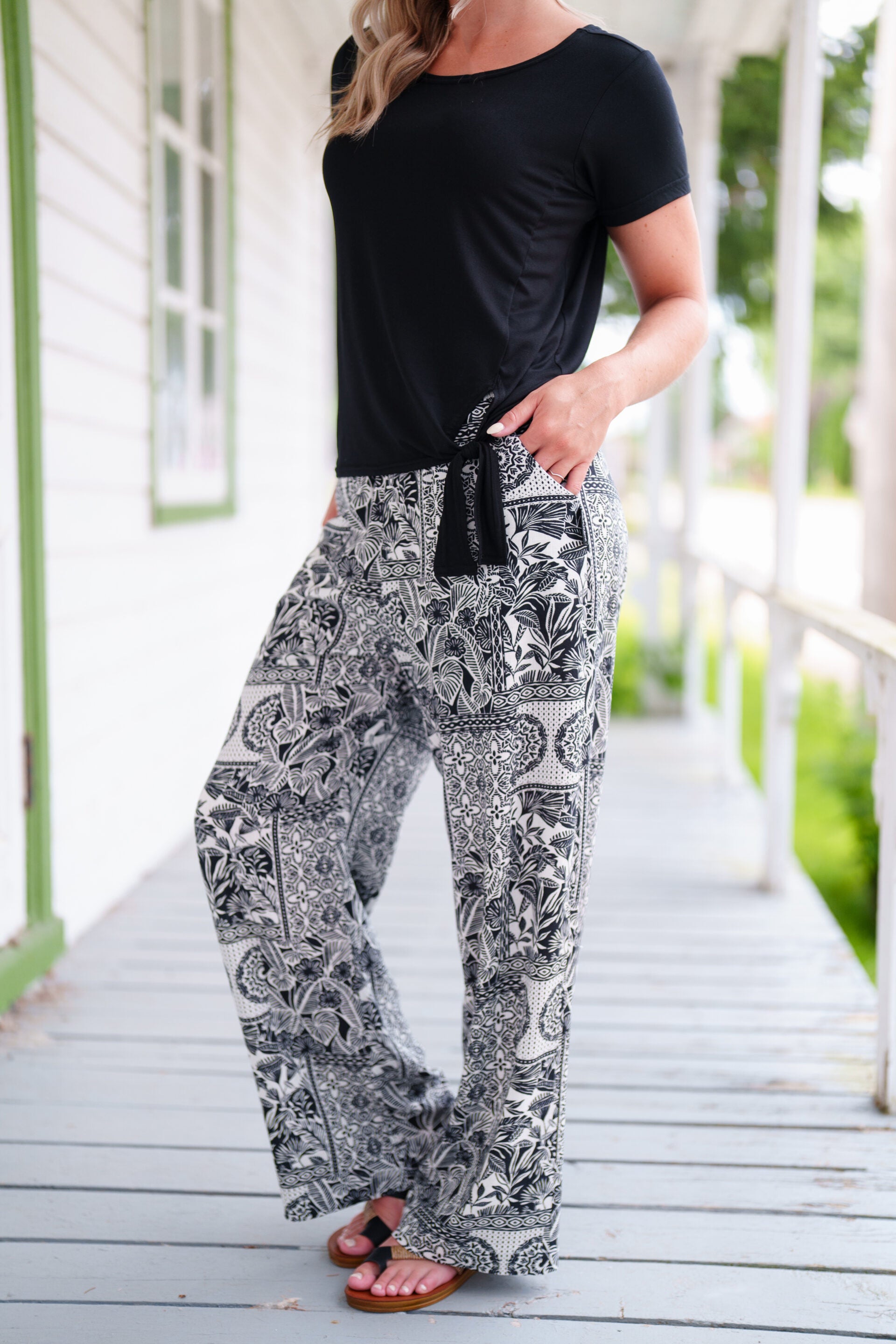 Neck down view of a woman wearing the Mango Pants from Rien ne se Perd in Black/Ivory tropical print, palazzo pants with an elastic waist and pockets. She is wearing them with a black top and standing on a porch. 
