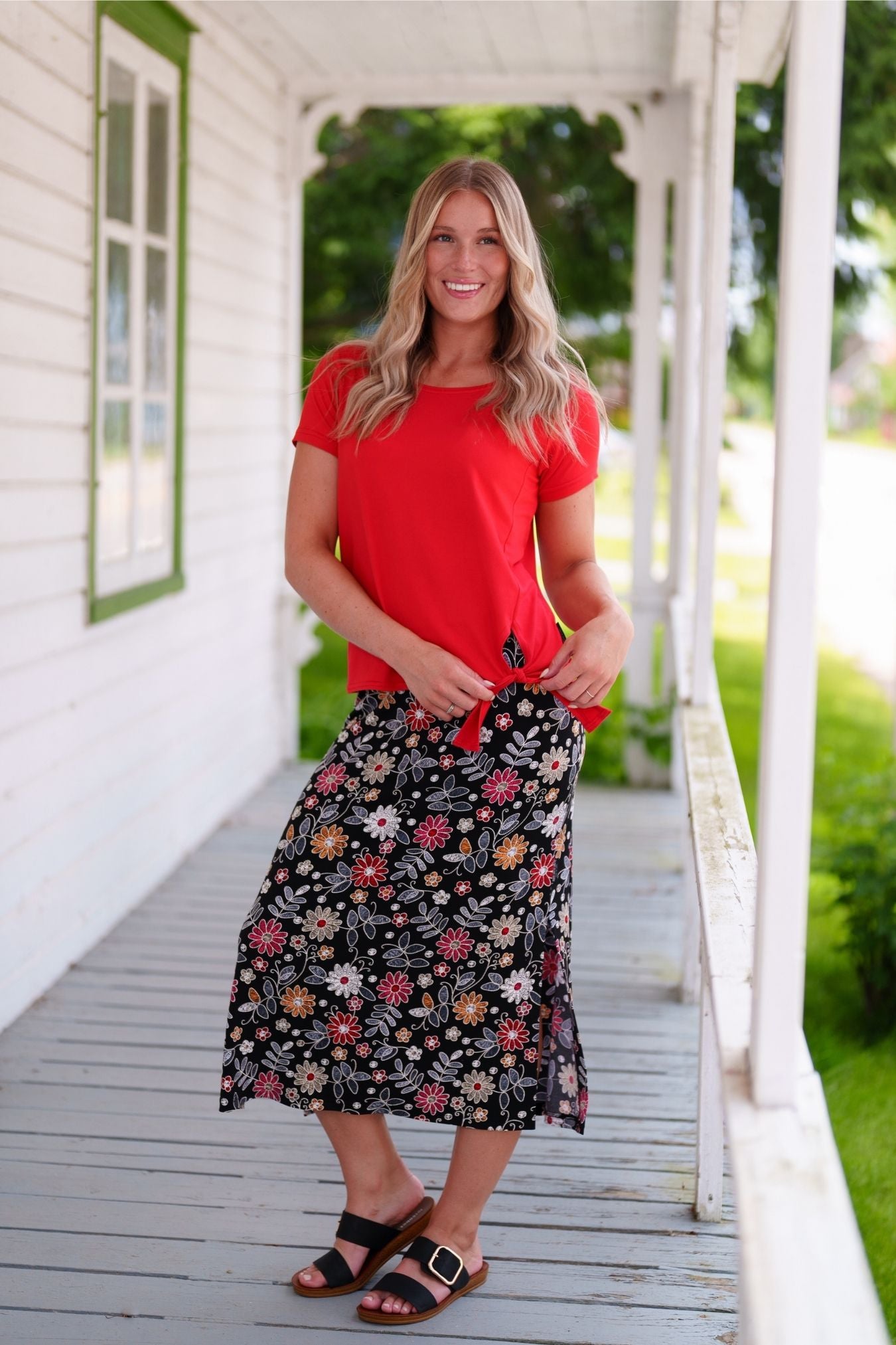 A woman wearing the Fiesta 7/8 Skirt by Rien ne se Perd in Colourful Daisies, a slightly A-line skirt with an elastic waist and side slits. She is wearing it with a red top and standing on a wooden porch.