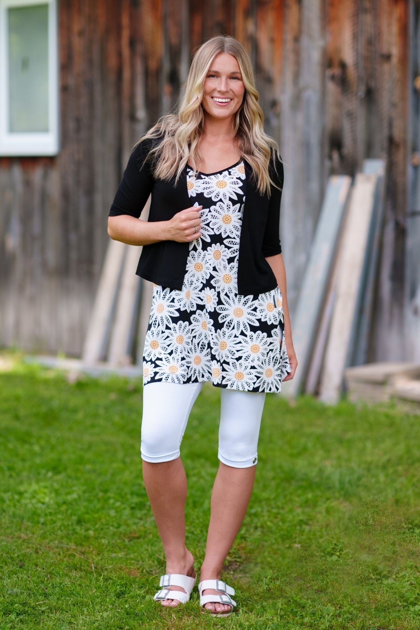 A woman wearing the Whisky Tunic by Rien ne se Perd, a sleeveless A-line piece embroidered with daisies. The V-neckline is finished with bias tape. She is wearing it with white 3/4 leggings and a black bolero and standing outside by a wooden building. 