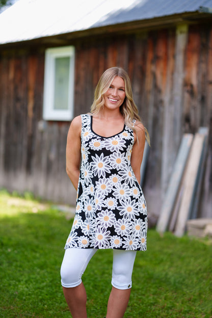 A woman wearing the Whisky Tunic by Rien ne se Perd, a sleeveless A-line piece embroidered with daisies. The V-neckline is finished with bias tape. She is wearing it with white 3/4 leggings and standing outside by a wooden building. 