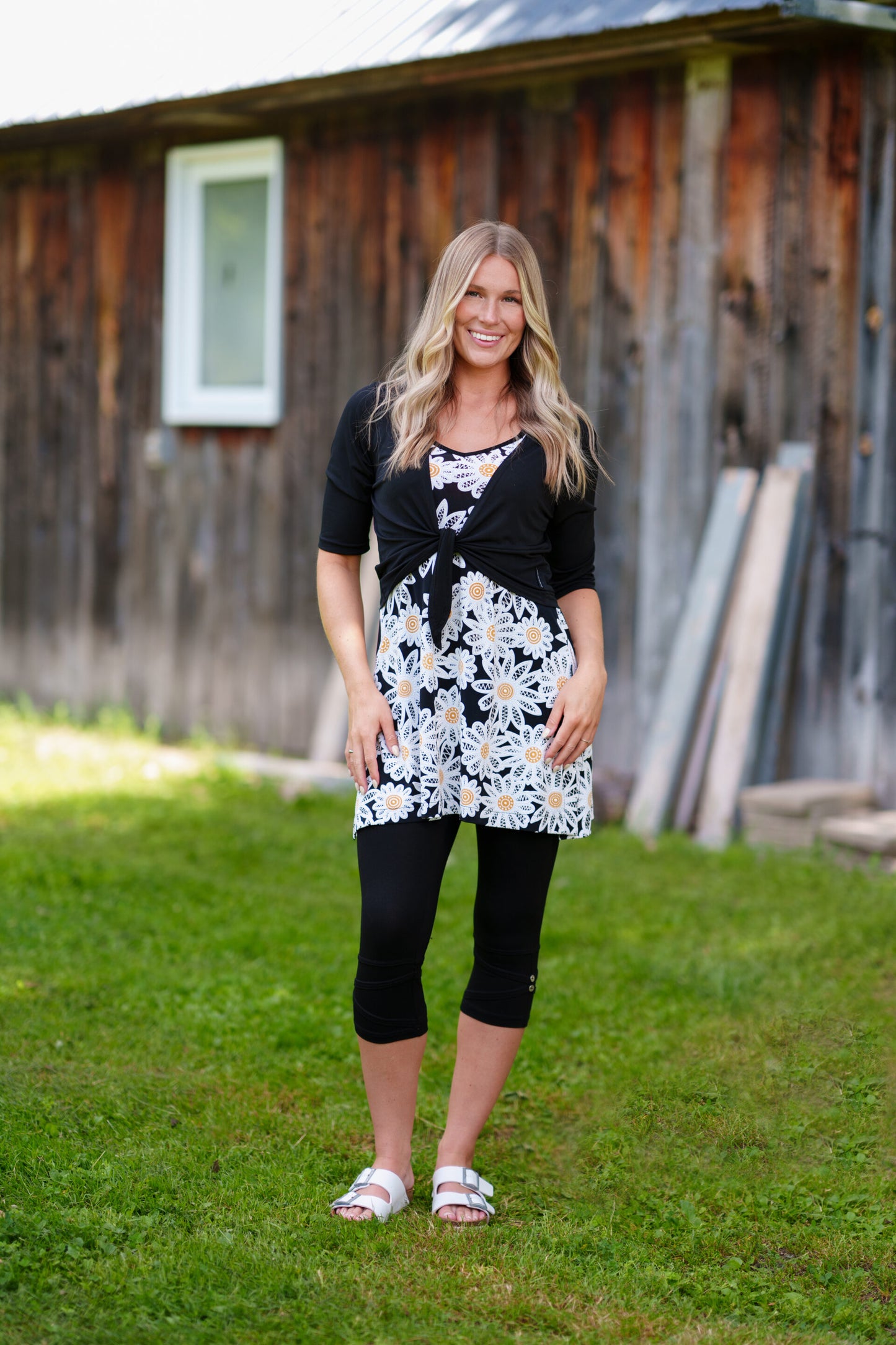 A woman wearing the Whisky Tunic by Rien ne se Perd, a sleeveless A-line piece embroidered with daisies. The V-neckline is finished with bias tape. She is wearing it with black 3/4 leggings and a black bolero and standing outside by a wooden building. 