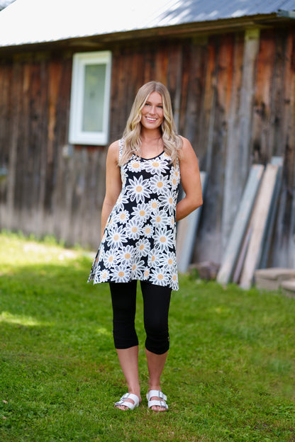 A woman wearing the Whisky Tunic by Rien ne se Perd, a sleeveless A-line piece embroidered with daisies. The V-neckline is finished with bias tape. She is wearing it with black 3/4 leggings and standing outside by a wooden building. 
