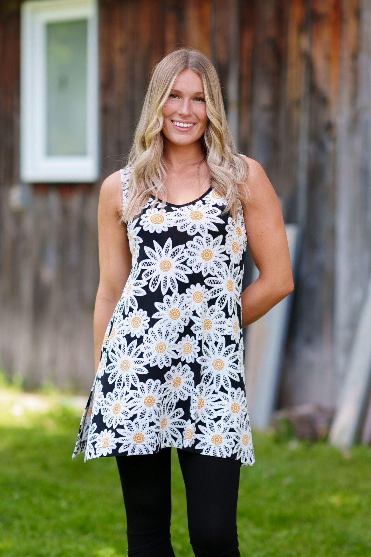 A woman wearing the Whisky Tunic by Rien ne se Perd, a sleeveless A-line piece embroidered with daisies. The V-neckline is finished with bias tape. She is wearing it with black leggings and standing outside by a wooden building. 